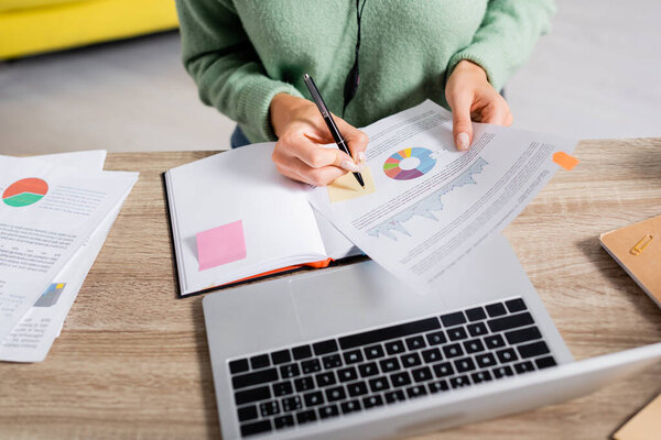 Cropped view of teleworker writing on sticky note on document with charts near notebook and laptop on blurred foreground 