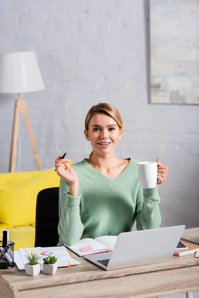 Smiling Teleworker Holding Pen Cup Laptop Documents Blurred Foreground Home — Stock Photo, Image