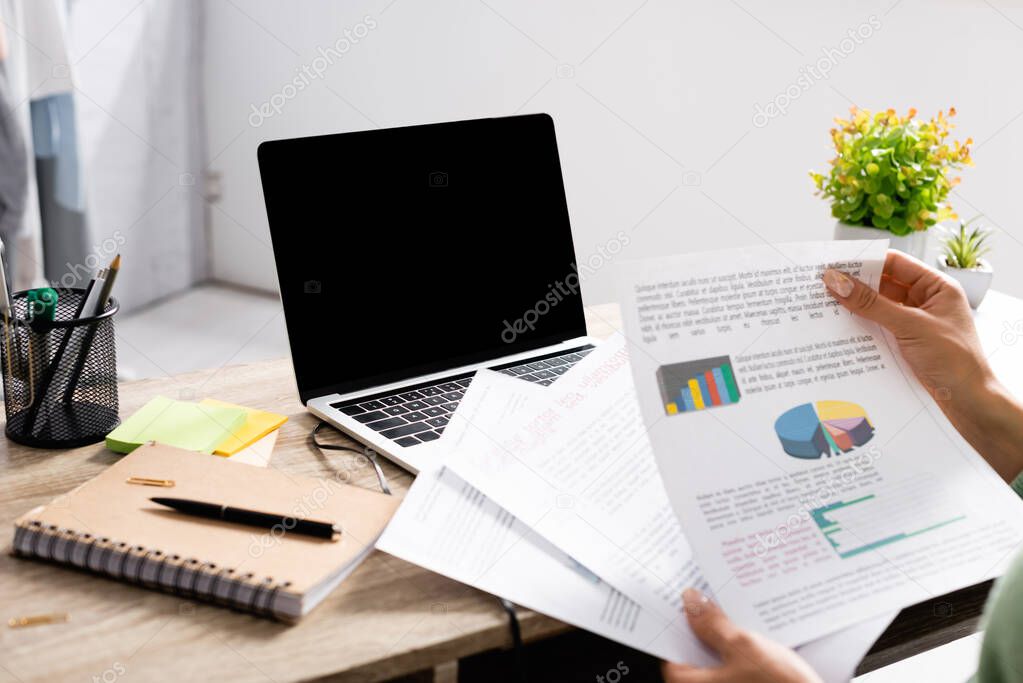 Cropped view of freelancer holding papers with charts near laptop with blank screen and notebook on table 