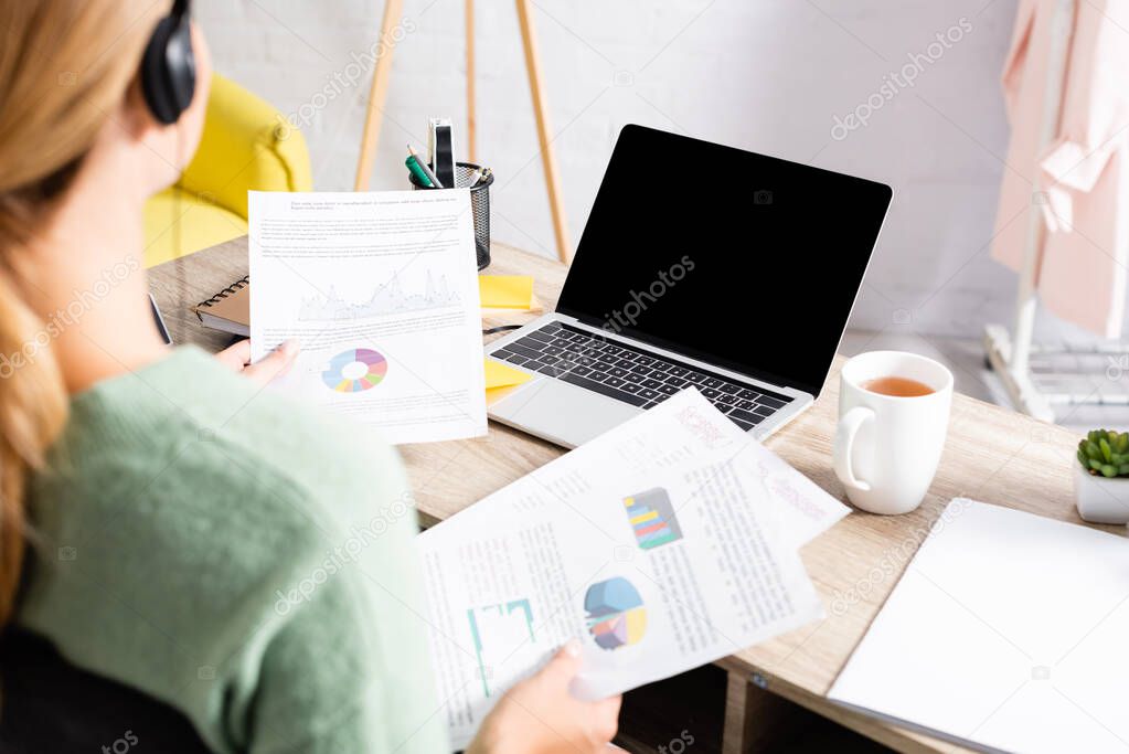 Laptop with blank screen near cup of tea and freelancer in headset holding documents on blurred foreground 