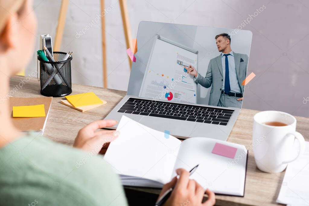 Businessman pointing at charts on flipchart on screen of laptop near freelancer holding pen and notebook on blurred foreground during webinar 