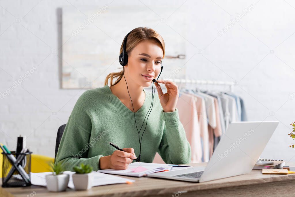 Smiling freelancer using headset and holding pen near notebook during video chat on laptop on blurred foreground 
