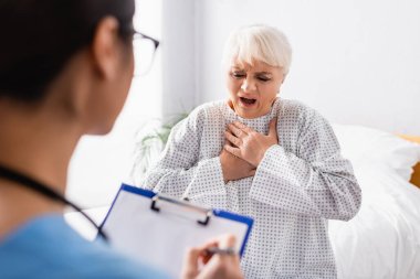 senior woman touching chest while suffering from asthma attack near nurse writing on clipboard on blurred foreground clipart