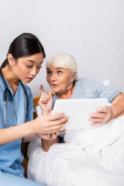 Heyecanlı yaşlı kadın dijital tablet kullanırken şaşırmış Asyalı hemşireyle fikir jesti yapıyor. 