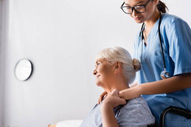 smiling senior woman holding hand of young asian nurse while looking away in hospital clipart