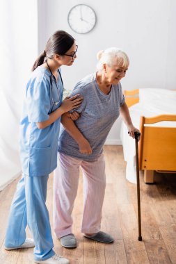 Asyalı hemşire hastanede çubukla yürüyen yaşlı kadına yardım ediyor.