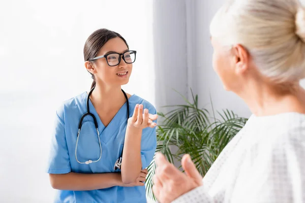 smiling asian nurse in eyeglasses gesturing while talking to senior woman in hospital