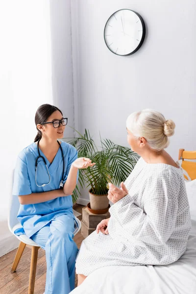 Young Asian Nurse Gesturing While Sitting Talking Senior Woman Hospital — Stock Photo, Image