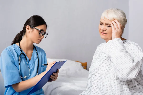 Asiatico Infermiera Occhiali Scrittura Appunti Vicino Invecchiato Donna Toccare Testa — Foto Stock