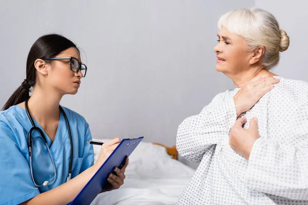 Young Asian Nurse Writing Clipboard Senior Woman Suffering Pain Shoulder — Stock Photo, Image