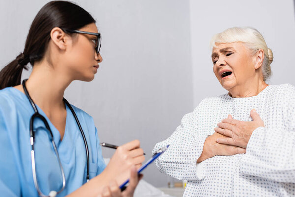 пожилая женщина трогает грудь, когда страдает от приступа астмы рядом с азиатской медсестрой, пишущей на планшете, размытый передний план