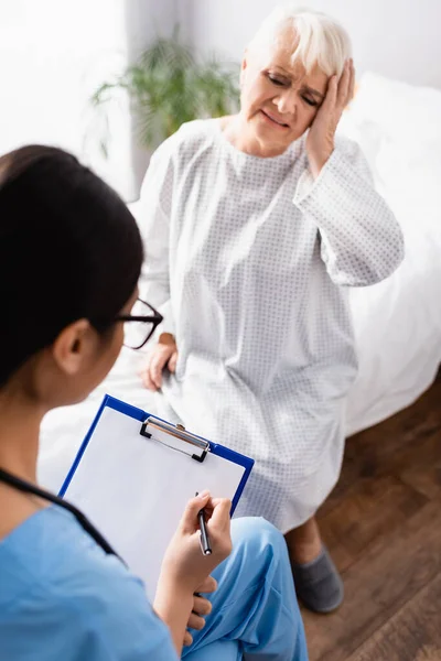 Young Nurse Writing Clipboard Senior Woman Touching Head While Suffering — Stock Photo, Image