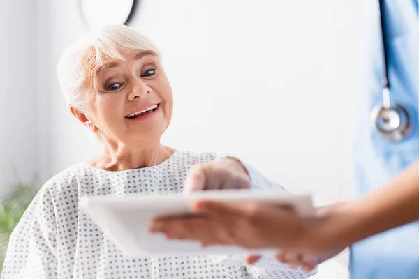 happy aged woman pointing at digital tablet in hands of nurse, blurred foreground