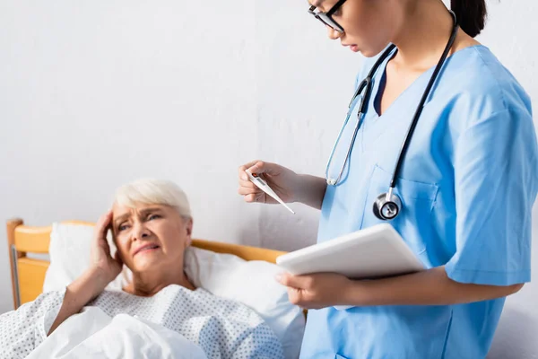 upset asian nurse with digital tablet looking at thermometer near ill senior woman suffering from headache on blurred background