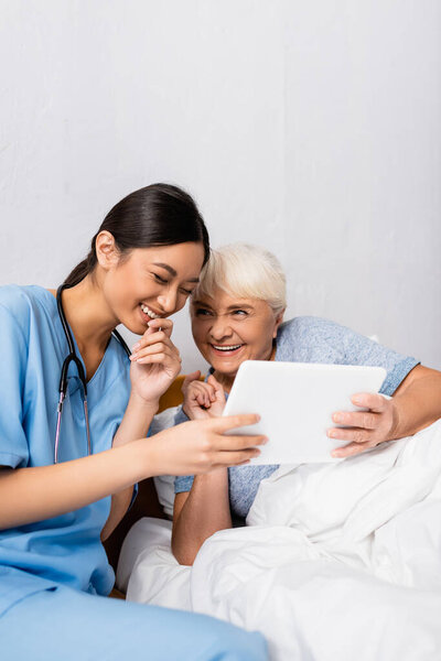 взволнованная азиатская медсестра и пожилая женщина смеются во время использования цифрового планшета вместе