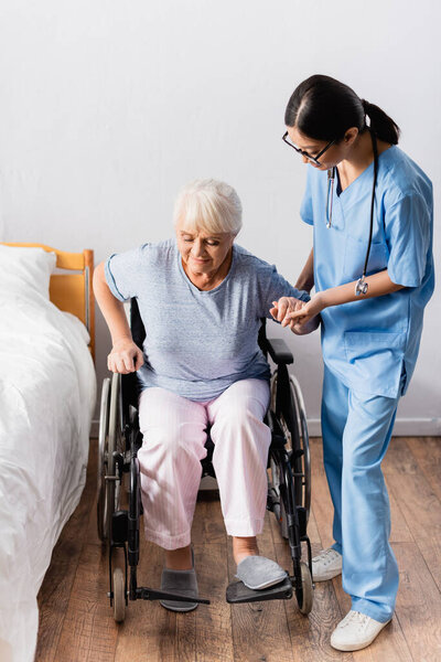 молодая азиатка, поддерживающая пожилую женщину, встающую с инвалидного кресла