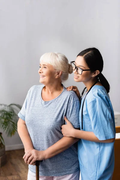 Lycklig Äldre Kvinna Med Käpp Och Ung Asiatisk Sjuksköterska Ler — Stockfoto