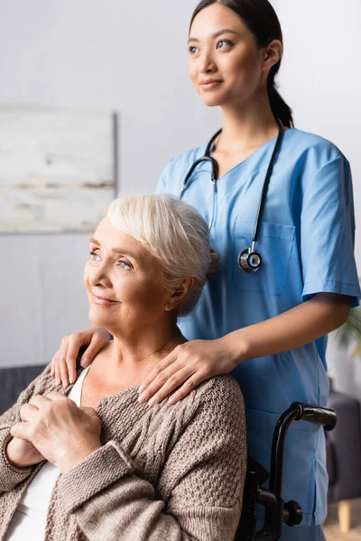 Joven Asiática Enfermera Tocando Hombros Sonriente Anciana Mirando Lejos Silla — Foto de Stock
