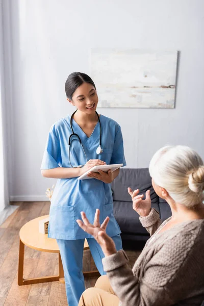 Anziana Donna Gesturing Mentre Parlando Con Sorridente Asiatico Infermiera Holding — Foto Stock