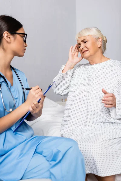 Young asian woman writing on clipboard near elderly woman suffering from headache — Stock Image Young Asian Woman Writing Clipboard Elderly Woman Suffering Headache Royalty Free Stock Photos
