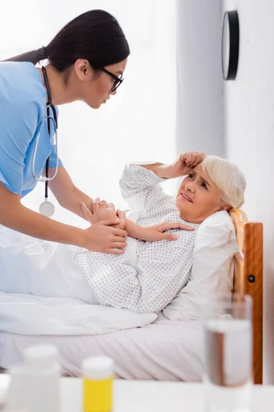 Young asian woman touching sick senior woman lying in bed and suffering from headache, blurred foreground — Stock Image Young Asian Woman Touching Sick Senior Woman Lying Bed Suffering Royalty Free Stock Photos