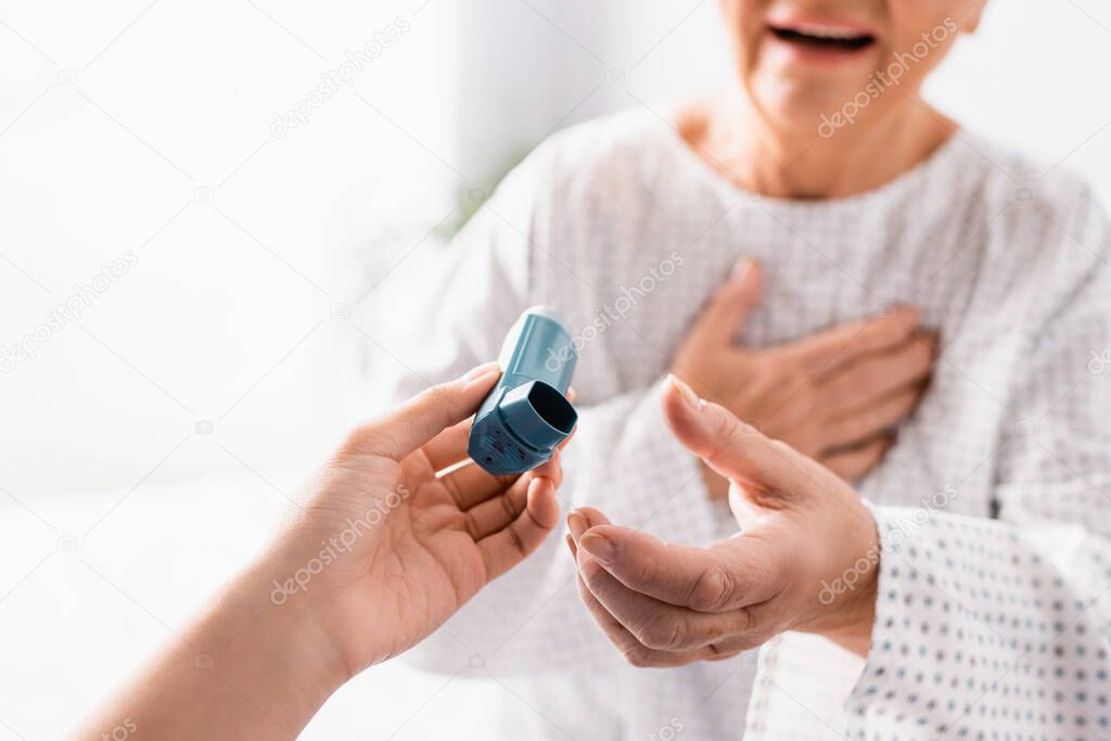 cropped view of nurse giving inhaler to aged woman suffering from asthma attack on blurred background