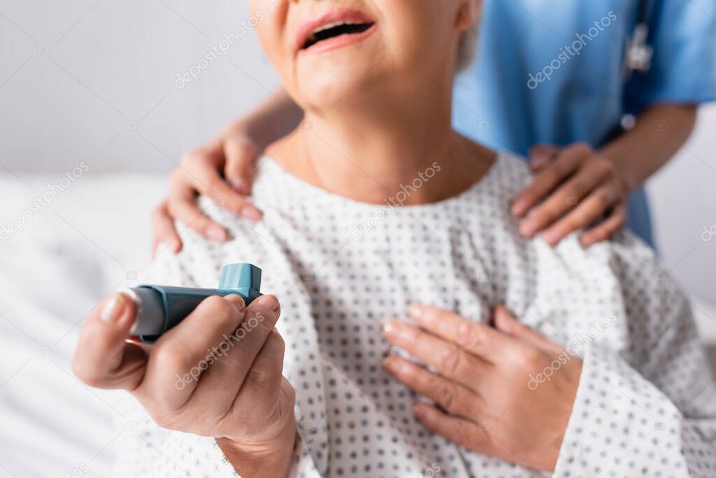 cropped view of elderly woman holding inhaler while suffering from asthma attack near nurse touching her shoulders, blurred background