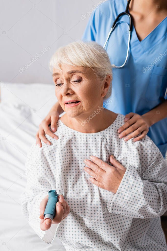 nurse touching shoulders of aged woman holding inhaler while suffering from asthma attack