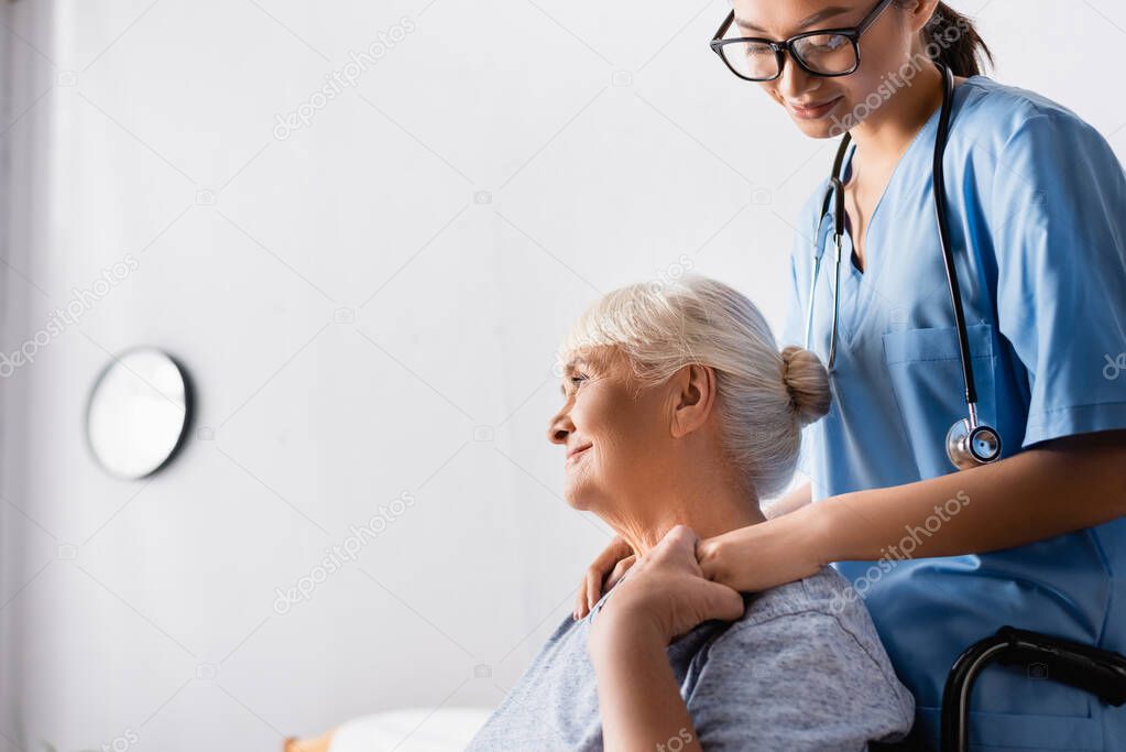 smiling senior woman holding hand of young asian nurse while looking away in hospital