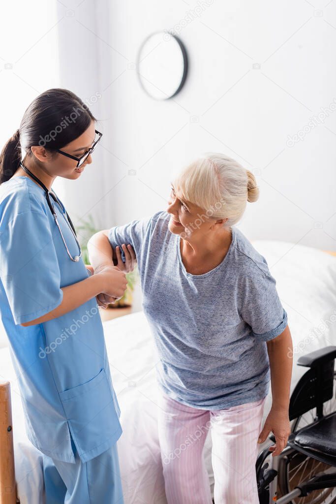 young asian nurse helping smiling, senior woman getting up from wheelchair