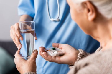 partial view of nurse giving pills and glass of water to elderly woman, selective focus clipart