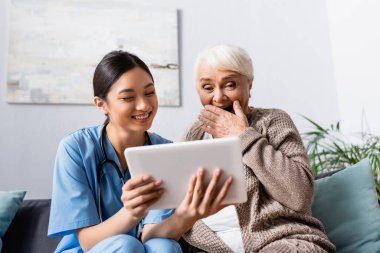 laughing senior woman covering mouth with hand while looking at digital tablet in hands of asian nurse clipart