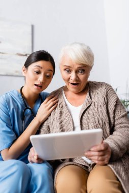 surprised asian woman touching shoulder of amazed senior woman looking at digital tablet clipart