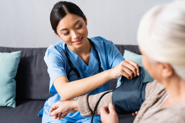 young asian nurse fixing cuff of tonometer on arm of aged woman in hospital