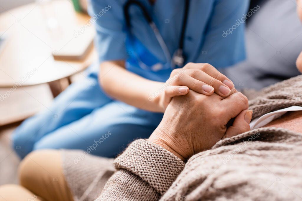 partial view of nurse touching hands of aged patient in nursing home, blurred background