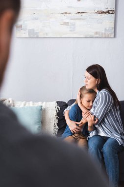 Depresyondaki kadın koltukta kucak dolusu çocuk kocasının yanında bulanık ön planda. 