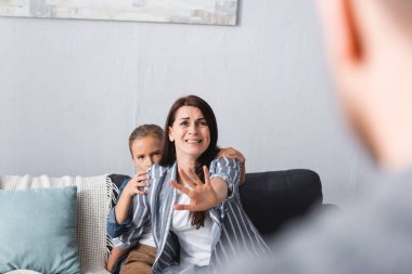 Scared woman showing stop gesture near child and abusive husband on blurred foreground 