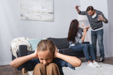 Kid covering ears while father quarrelling at mother on couch on blurred background at home  clipart