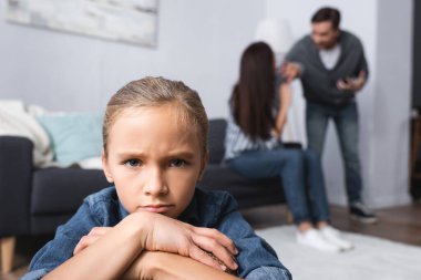 Sad girl looking at camera while parents quarrelling on blurred background  clipart
