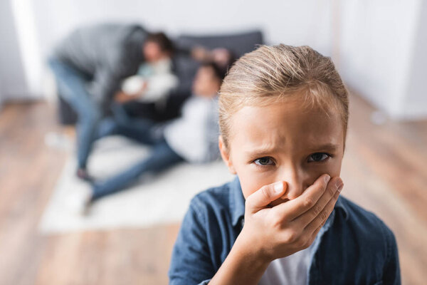 Offended child covering mouth with hand while parents fighting on blurred background in living room 