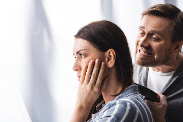 Woman with hand near cheek looking away beside irritated husband with waist belt at home 