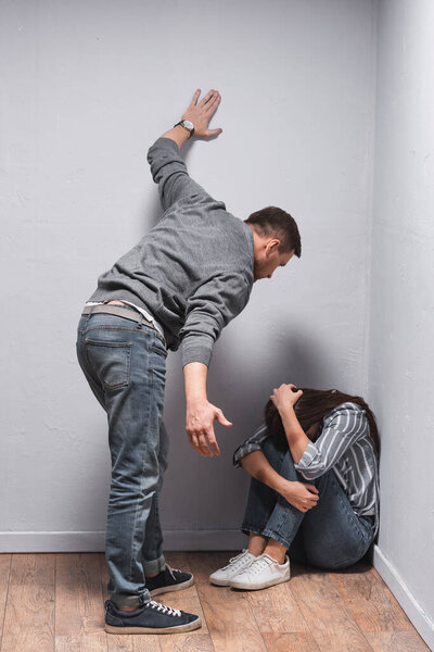 Abusive husband standing near wife with bruises sitting on floor near walls 