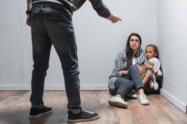 Child with soft toy and woman with bruises sitting on floor near abusive husband pointing with finger on blurred foreground  clipart