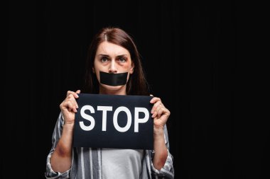 Victim of domestic abuse with bruises and adhesive tape on mouth holding card with stop lettering isolated on black  clipart