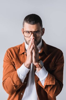 Gözlüklü şık bir adam. Dua eden elleri var. Gri renkli kameraya bakıyor. 