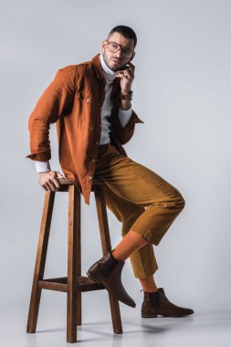 Terracotta ceketli ve kahverengi ayakkabılı şık bir adam gri arka planda ahşap sandalyenin yanında poz veriyor. 