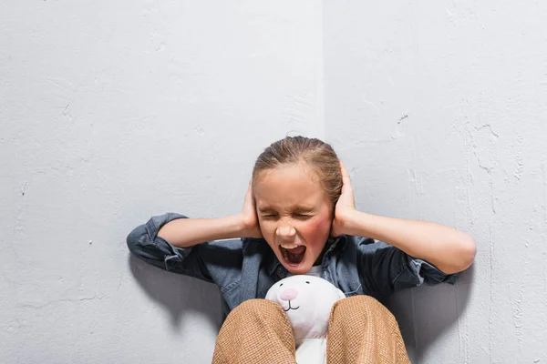 Menina Gritando Com Hematoma Rosto Cobrindo Orelhas Perto Brinquedo Macio — Fotografia de Stock