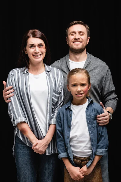 Smiling man hugging unhappy wife and daughter with bruises isolated on black