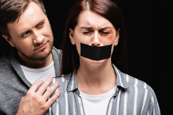 Abusive man hugging offended wife with bruise and adhesive tape on mouth isolated on black 