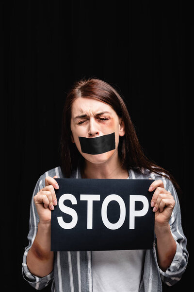 Victim of domestic violence with adhesive tape on mouth holding card with stop lettering isolated on black 